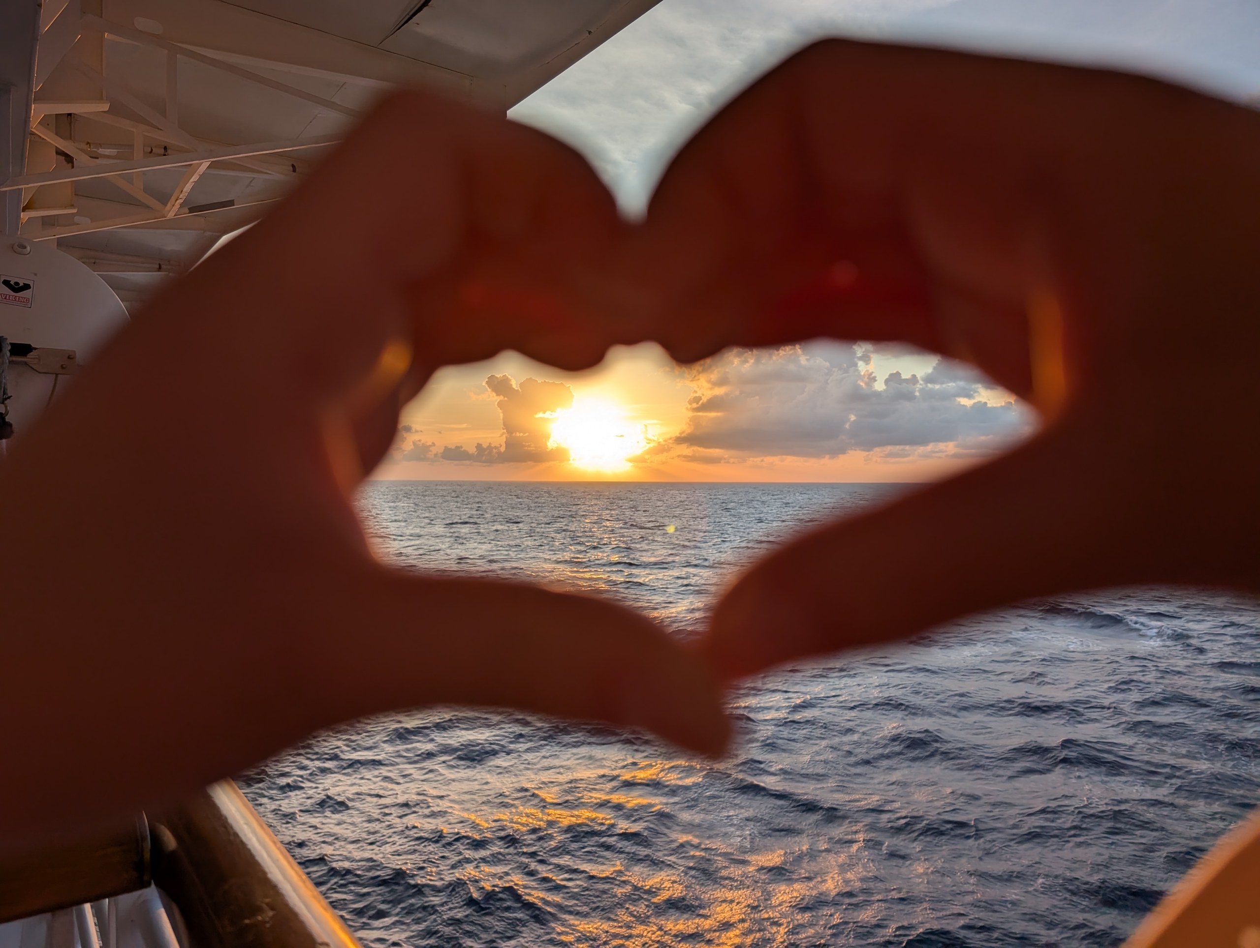 Two hands forming a heart shape in front of the sun setting over the Caribbean, viewed from a Harmony of the Seas stateroom balcony.