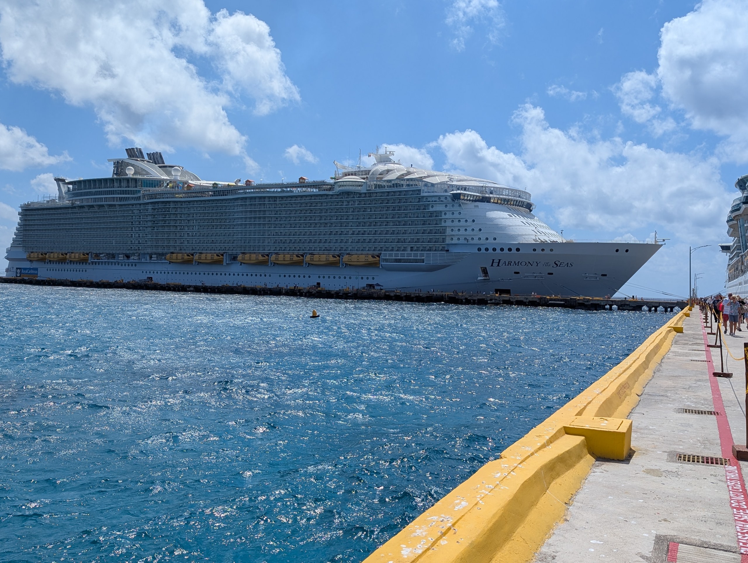 Royal Caribbean's Harmony of the Seas docked alongside a tropical port pier under a bright blue sky.