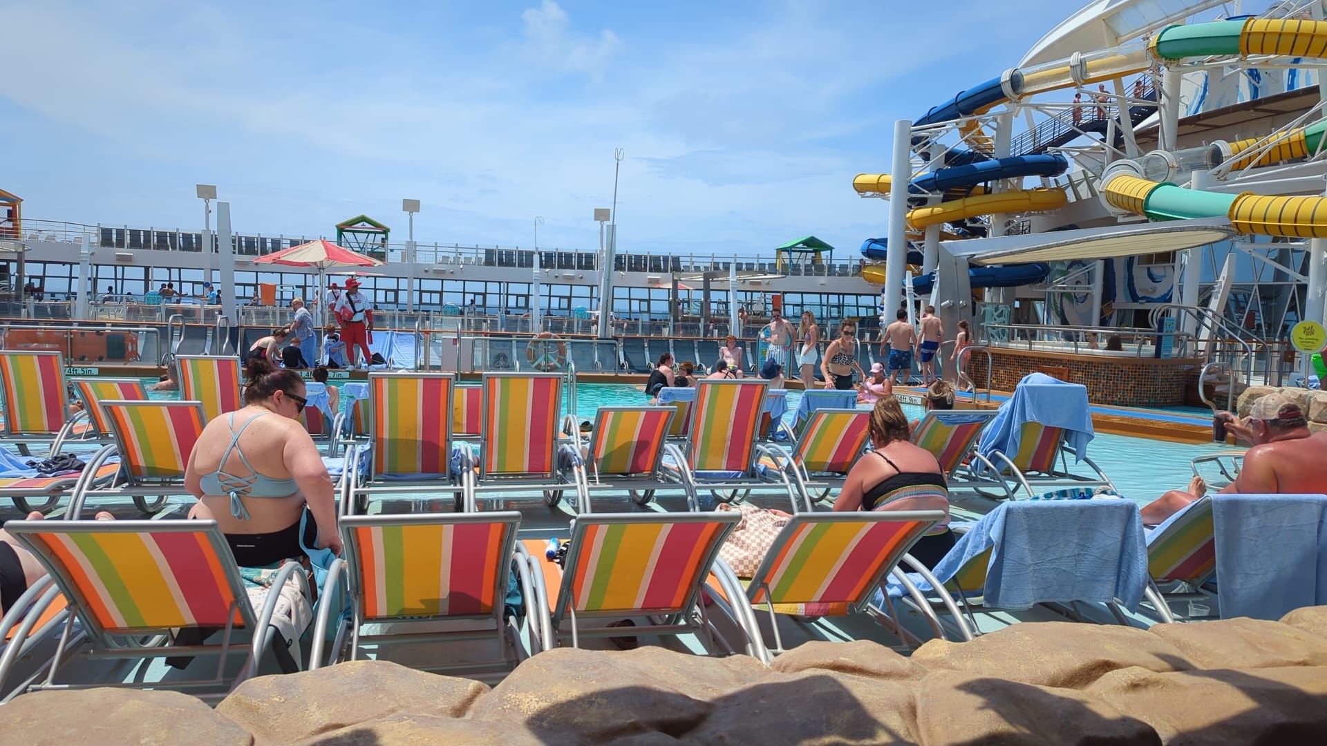 Crowded pool deck on Harmony of the Seas with rainbow-striped lounge chairs and the colorful Perfect Storm waterslides in the background.