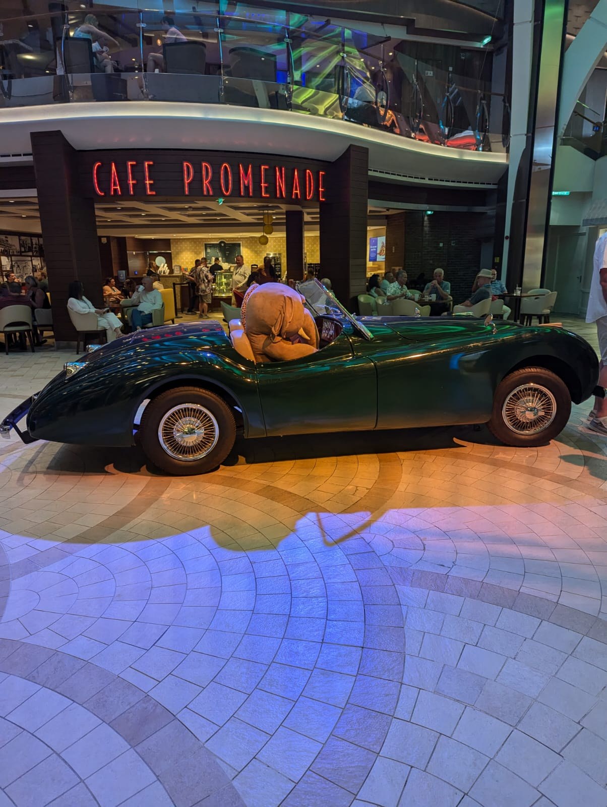The Royal Promenade aboard Harmony of the Seas with a vintage British roadster on display in front of the Cafe Promenade signage.
