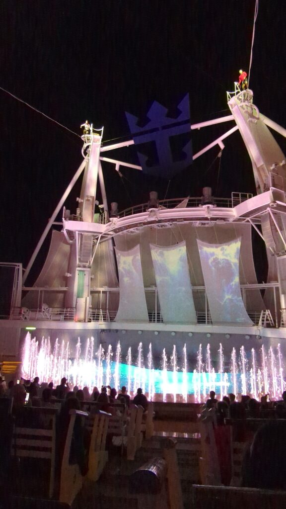 Illuminated AquaTheater stage at the back of Symphony of the Seas at night with performers in costume and the Royal Caribbean crown and anchor logo lit on the deck above.