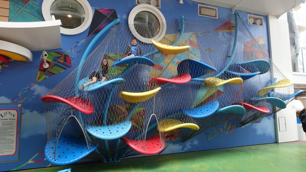 Two kids climbing the colorful kite-themed climbing net wall in the Boardwalk neighborhood on Symphony of the Seas.