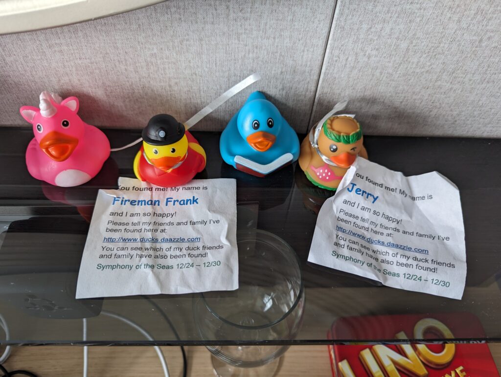 Four hidden rubber ducks lined up in a stateroom on Symphony of the Seas, with the printed cards from the cruise-ducking tradition explaining the tags Fireman Frank and Jerry, dated 12/24 to 12/30.