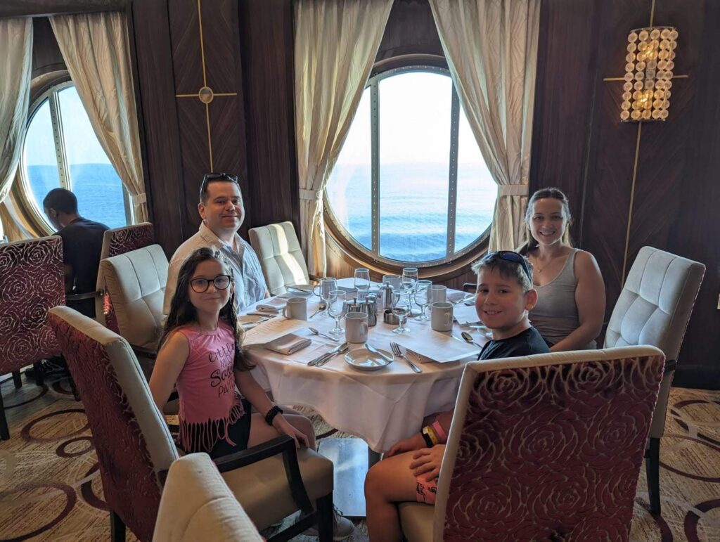 Alex, Vika, Josh, and Emily at a round dining table on Symphony of the Seas with a large oval porthole window behind them showing the ocean, formal place settings on the table.