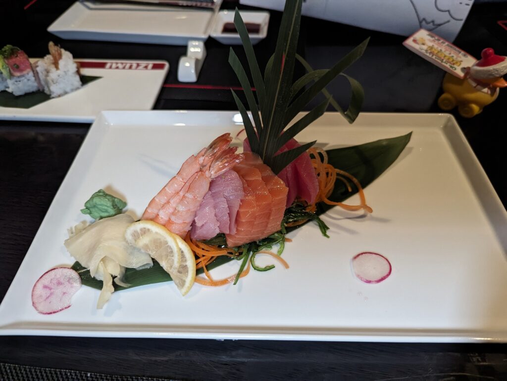 A composed sashimi platter on a long white plate with shrimp, salmon, tuna, and yellowtail arranged with carrot ribbons, lemon, ginger, and a leaf garnish at Izumi on Symphony of the Seas.