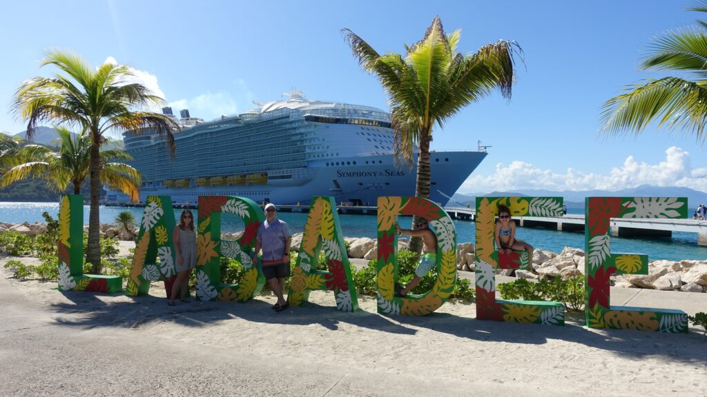 The Ostrovsky family standing at the colorful Labadee welcome sign on Royal Caribbean's private Haitian destination, with Symphony of the Seas docked in the background.