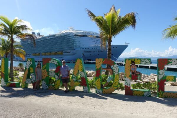 The Ostrovsky family standing at the colorful Labadee welcome sign on Royal Caribbean's private Haitian destination, with Symphony of the Seas docked in the background.