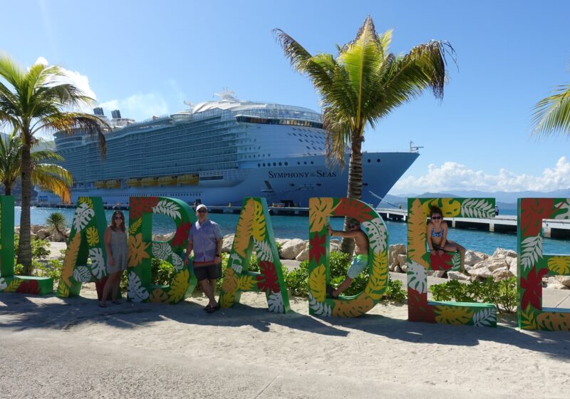 The Ostrovsky family standing at the colorful Labadee welcome sign on Royal Caribbean's private Haitian destination, with Symphony of the Seas docked in the background.