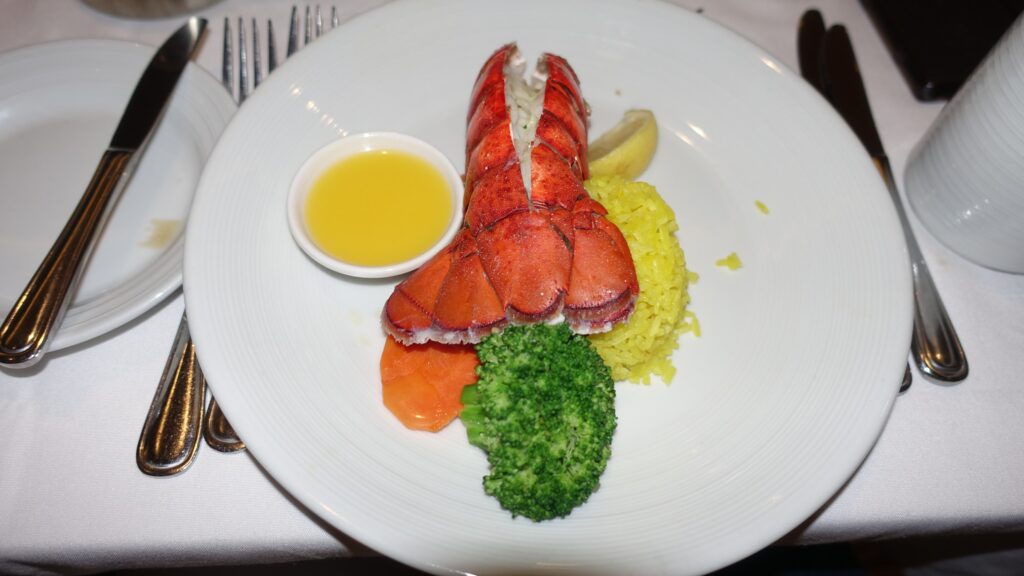 Plate from the main dining room on Symphony of the Seas with a whole grilled lobster tail in shell, drawn butter, yellow saffron rice, broccoli, and tomato.