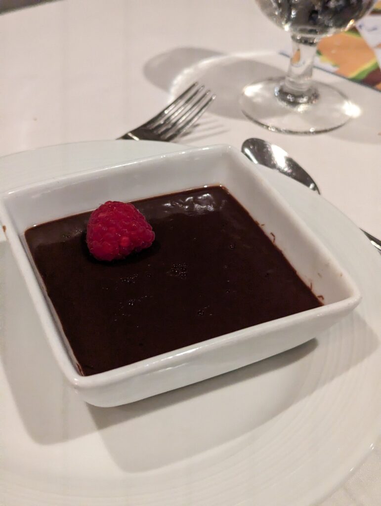 Square white ramekin of dark chocolate pot de crème topped with a single fresh raspberry, served on a white plate with a fork and spoon at the main dining room on Symphony of the Seas.