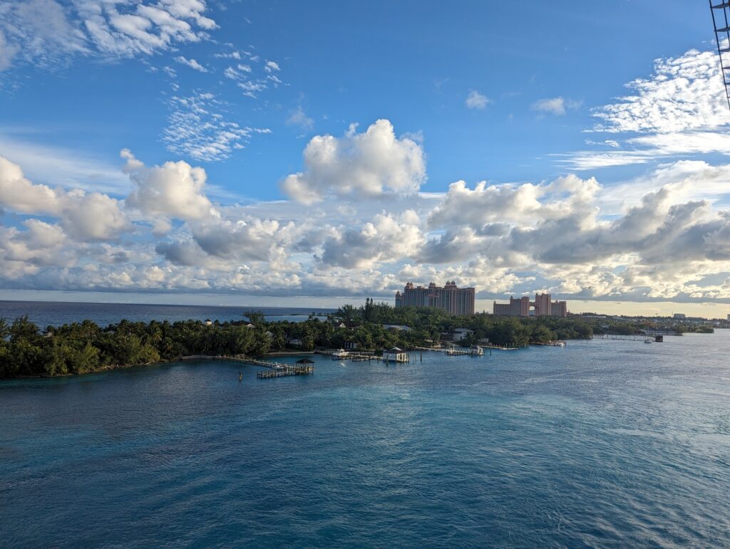 View from the deck of Symphony of the Seas approaching Nassau, Bahamas, with the pink towers of the Atlantis resort visible across the harbor under a partly cloudy sky.