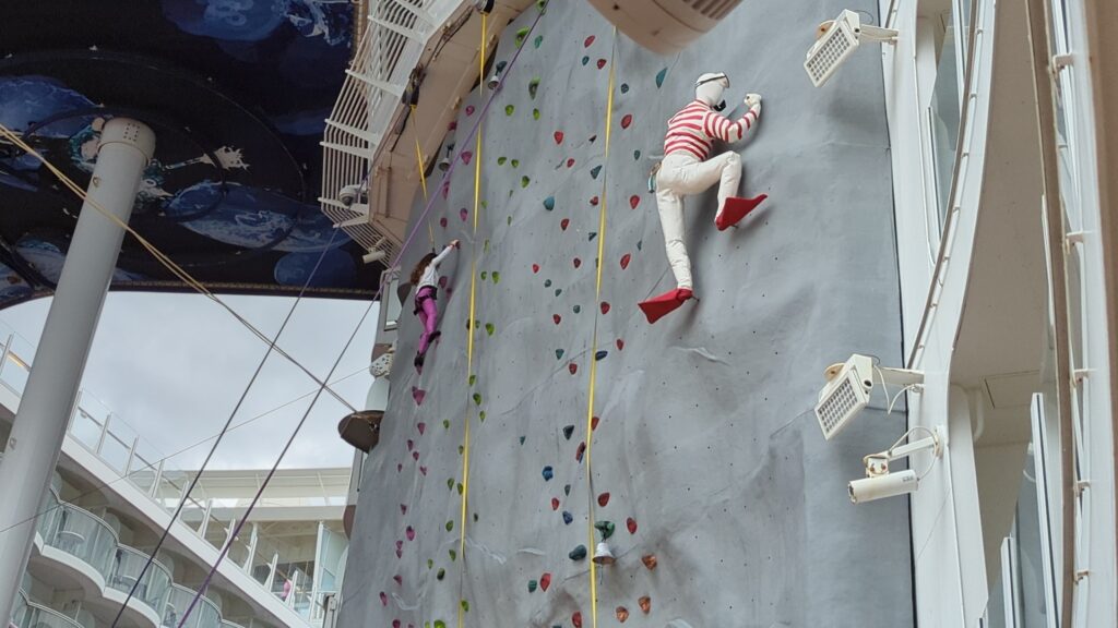 Two climbers on the outdoor rock climbing wall on Symphony of the Seas, one in pink and one in red and white stripes, with the ship's deck and stage shell visible behind them.