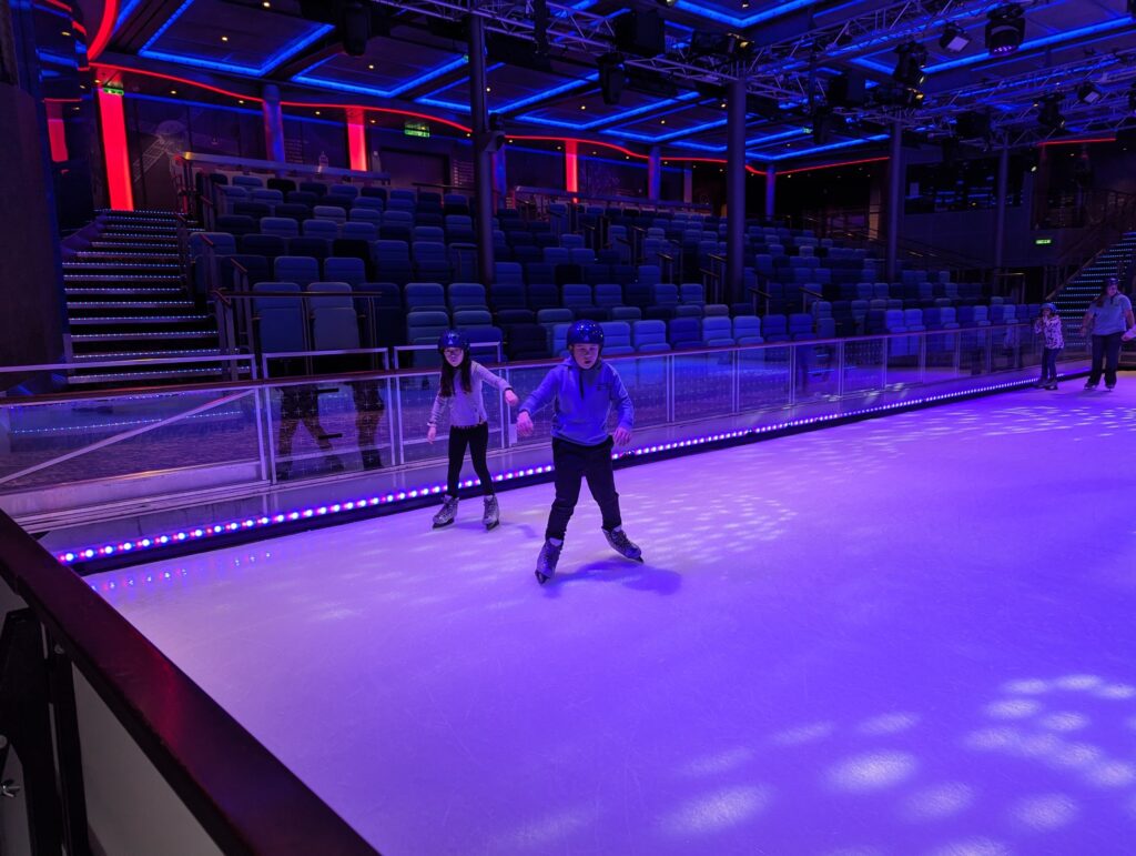 Josh and Emily in helmets ice skating on the Studio B rink on Symphony of the Seas, with purple and blue stage lighting and empty stadium seating around them.
