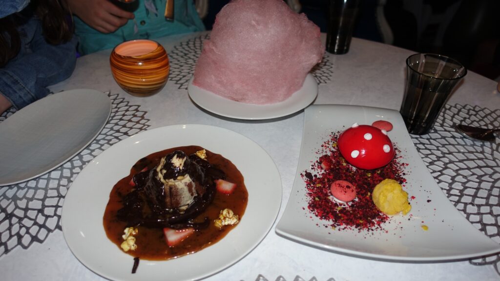 Wonderland dessert course on Symphony of the Seas with a chocolate-covered ice cream dome on a saucer, a pink cotton candy cloud on a plate, and a red-and-white mushroom-shaped dessert with macaron and crumb on a third plate.