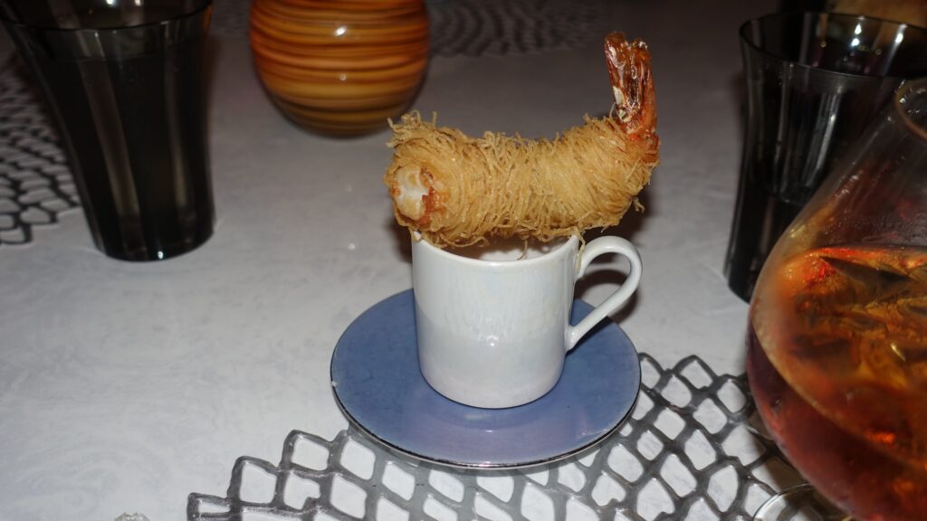 A single tempura-fried shrimp wrapped in fine kataifi noodle threads, perched dramatically on an espresso cup over a blue saucer at the Wonderland restaurant on Symphony of the Seas.
