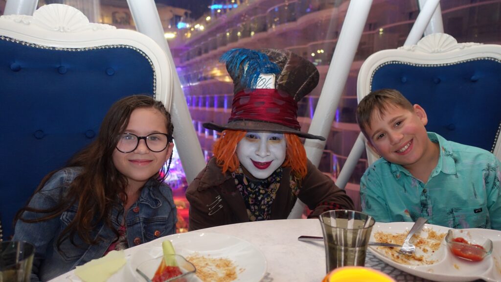 Emily and Josh seated at a Wonderland restaurant table on Symphony of the Seas with the Mad Hatter character in red wig and top hat between them, plates of dessert on the table.