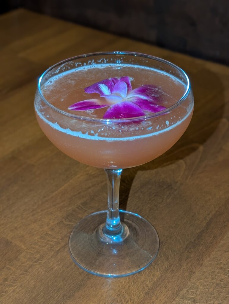 Ananda Spritz cocktail in a coupe glass garnished with a purple orchid, side angle