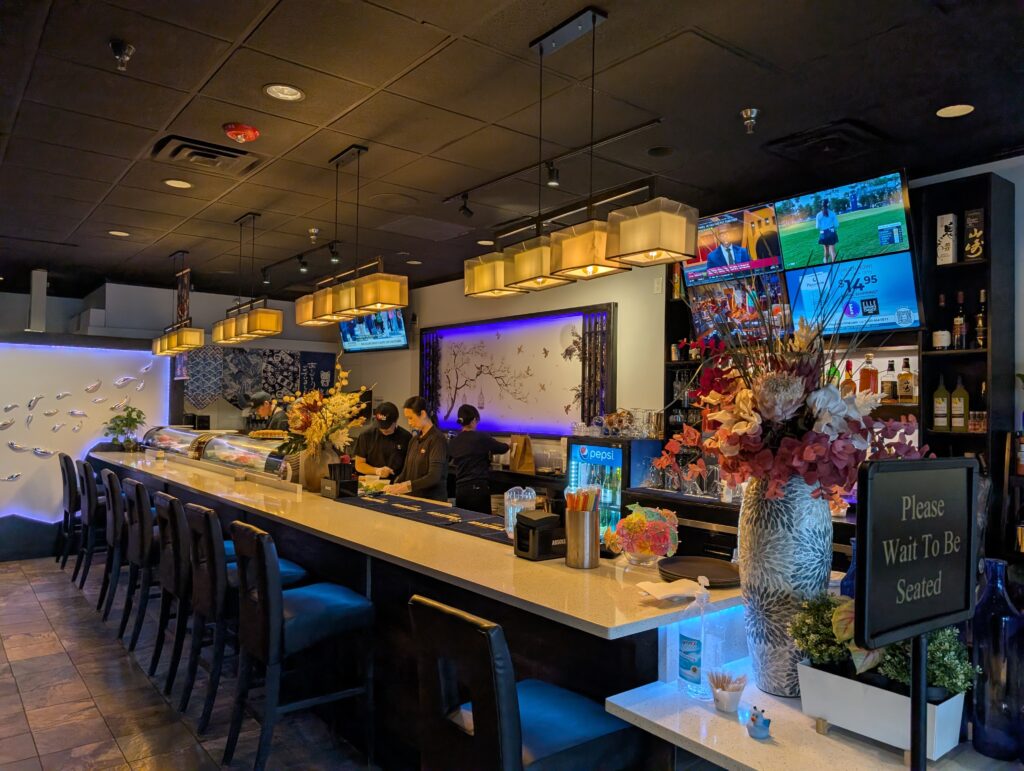 Sushi bar counter at Tekka Sushi with bar seating, pendant lights, and a Please Wait To Be Seated sign.