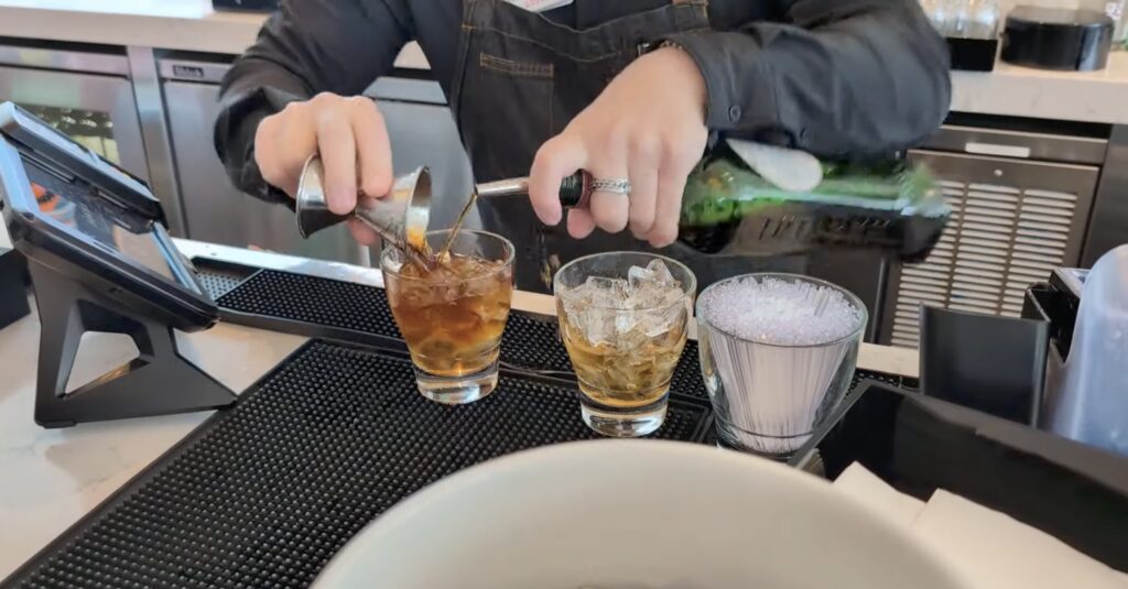 Bartender at the United Club by Gate C1 at Houston IAH pouring a freshly-shaken Manhattan into a rocks glass over ice, with a second glass and ice container on the bar mat.
