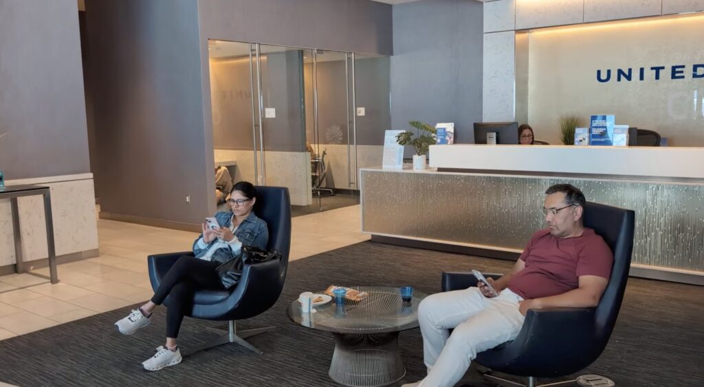 Front desk lobby of the United Club by Gate C1 at IAH with the United Club logo on the back wall, two travelers seated in modern black lounge chairs around a coffee table.
