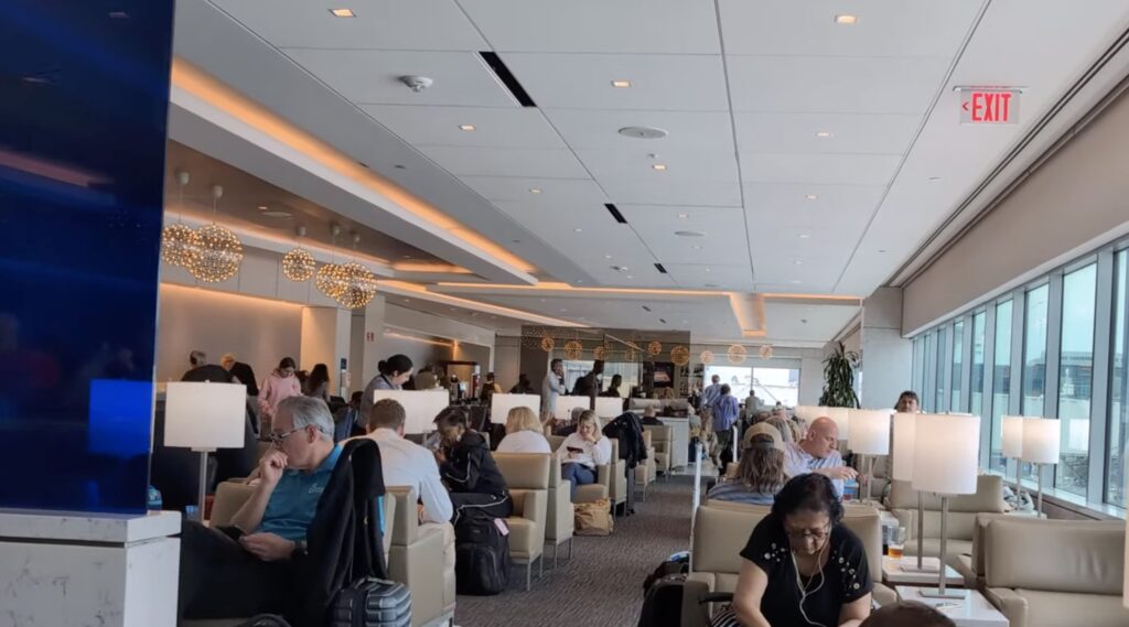 Wide view of the United Club by Gate C1 at IAH main lounge, with travelers seated in cream lounge chairs, modern pendant lights along the bar in the distance, and large windows on the right.