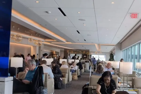 Wide view of the United Club by Gate C1 at IAH main lounge, with travelers seated in cream lounge chairs, modern pendant lights along the bar in the distance, and large windows on the right.