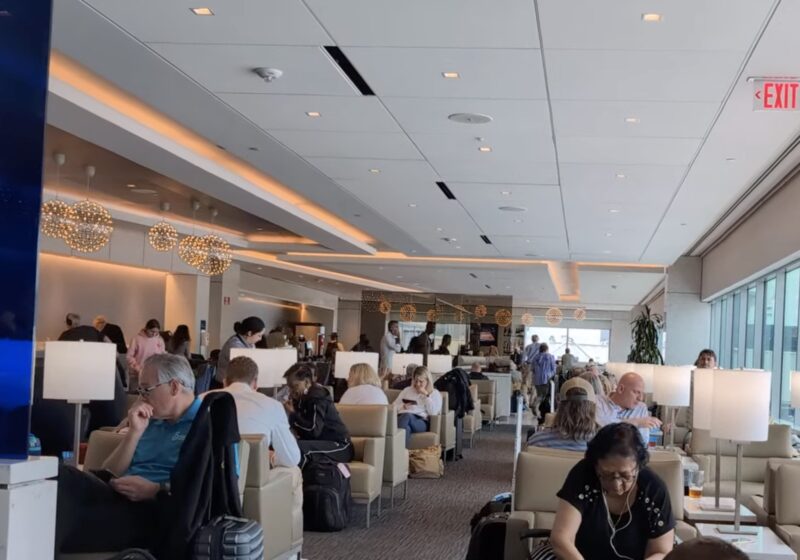Wide view of the United Club by Gate C1 at IAH main lounge, with travelers seated in cream lounge chairs, modern pendant lights along the bar in the distance, and large windows on the right.