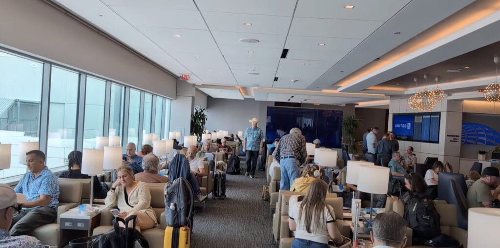 Main floor of the United Club by Gate C1 at IAH with floor-to-ceiling tarmac windows on the left, the United Club branded back wall in the center, and travelers seated in lounge chairs throughout.