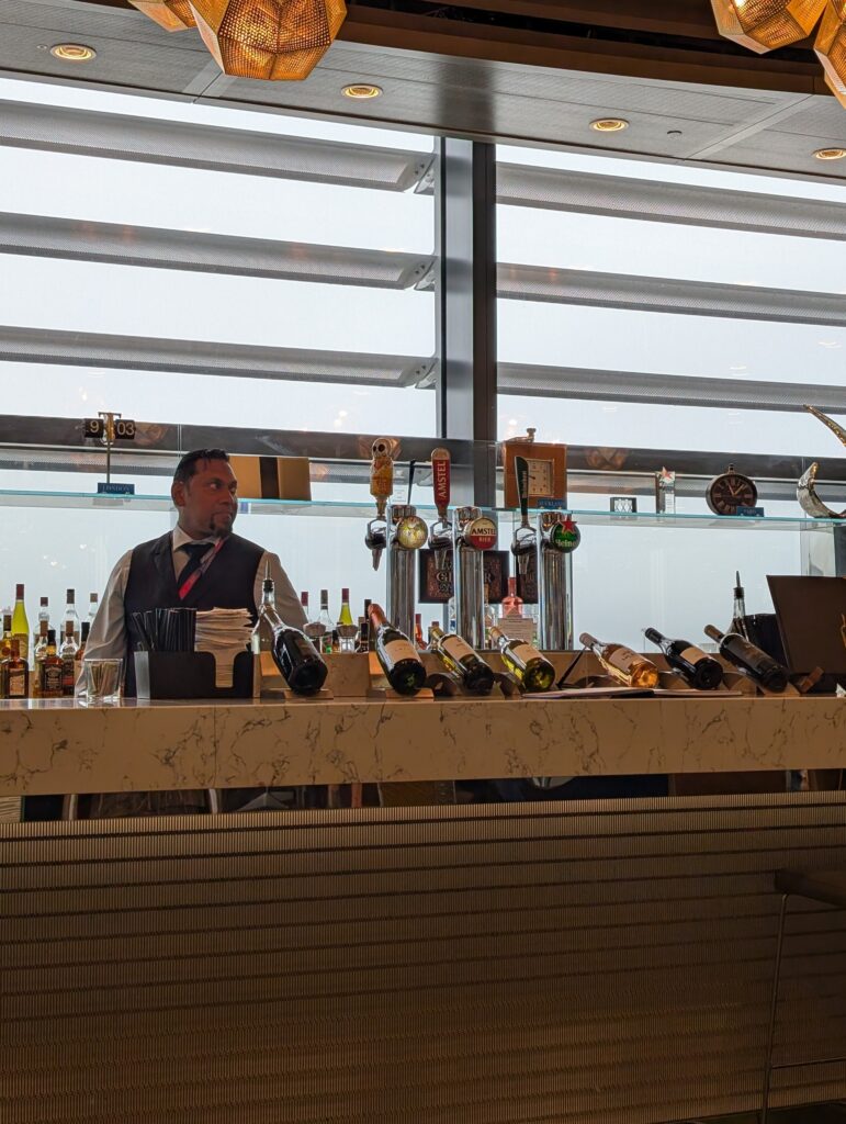 Bartender in a waistcoat behind the bar at the United Club at LHR Terminal 2, with a curated wine and beer selection lined up on the marble counter.
