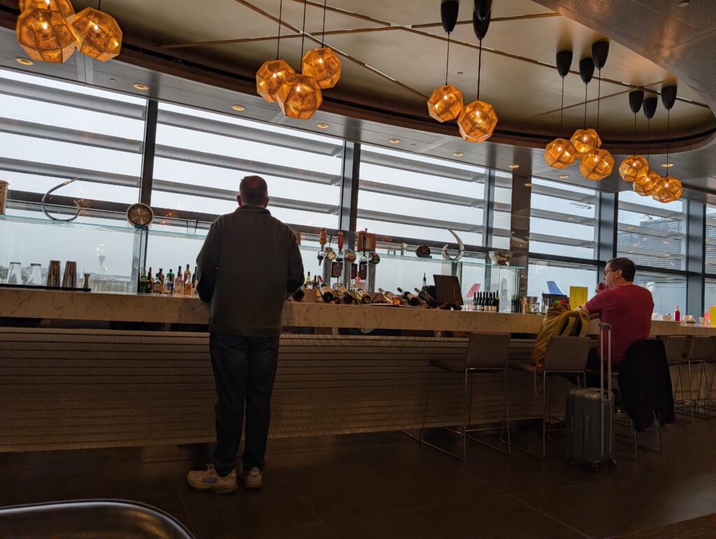 Wide view of the United Club bar at LHR Terminal 2 with iconic amber Tom Dixon-style pendant lights, a marble bar counter, patrons seated, and the tarmac visible through clerestory windows.