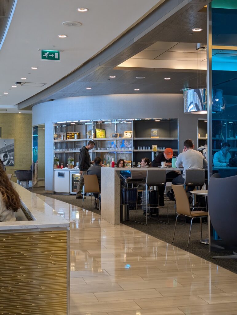 Self-serve buffet and beverage station at the United Club at LHR Terminal 2 with travelers plating food at a curved counter under bright recessed lighting.