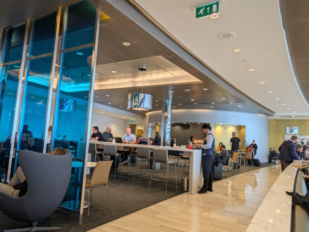 Communal dining area at the United Club at LHR Terminal 2 with blue back-lit glass walls, a flight information display, and a uniformed lounge attendant.