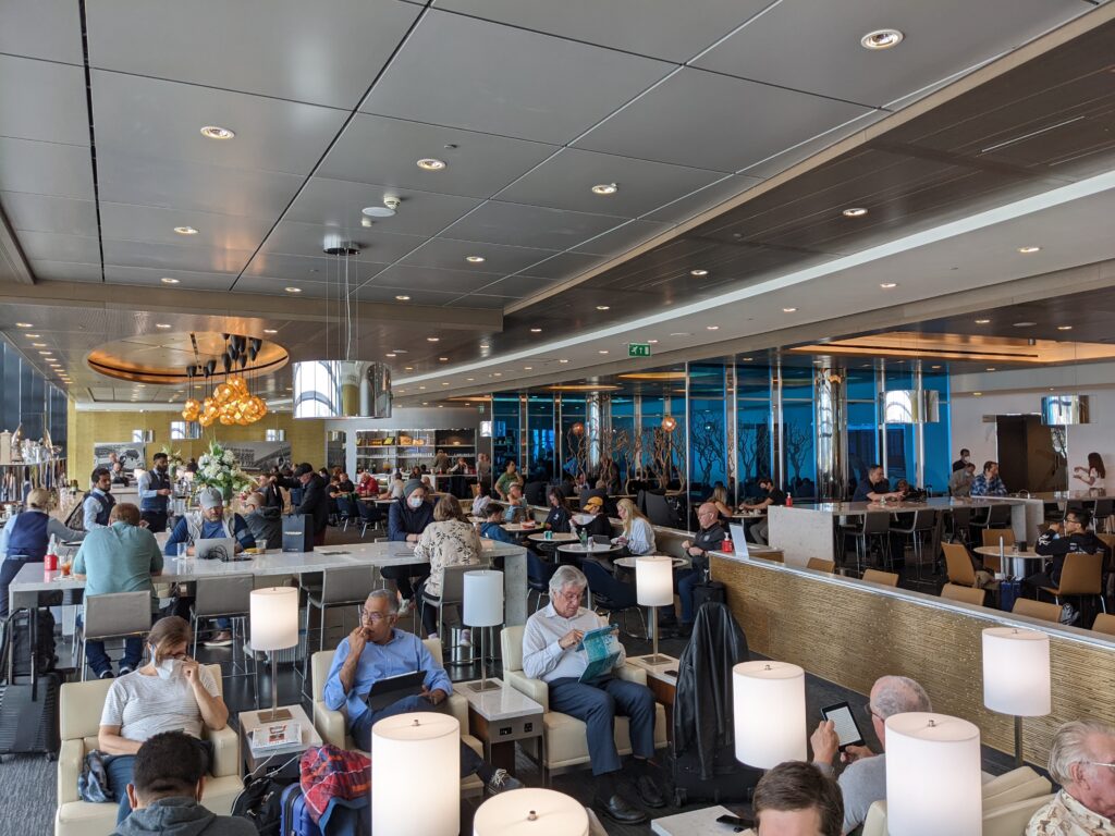 Wide view of the United Club main lounge at London Heathrow Terminal 2 with chandelier lighting, floor-to-ceiling windows, and lounge seating filled with travelers.
