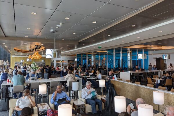Wide view of the United Club main lounge at London Heathrow Terminal 2 with chandelier lighting, floor-to-ceiling windows, and lounge seating filled with travelers.