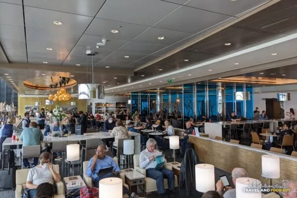 Wide view of the United Club main lounge at London Heathrow Terminal 2 with chandelier lighting, floor-to-ceiling windows, and lounge seating filled with travelers.
