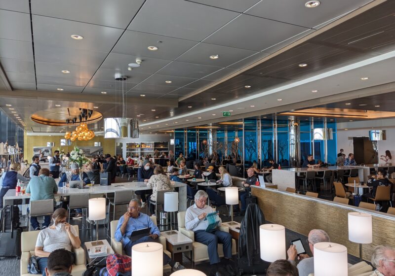Wide view of the United Club main lounge at London Heathrow Terminal 2 with chandelier lighting, floor-to-ceiling windows, and lounge seating filled with travelers.