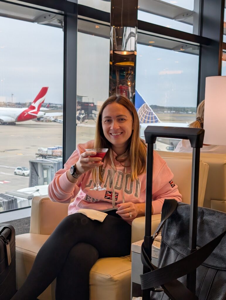 Vika in a pink London hoodie sitting in the United Club at LHR Terminal 2, holding a Manhattan cocktail with a Qantas A380 and a United Polaris-livery aircraft visible on the tarmac through floor-to-ceiling windows.