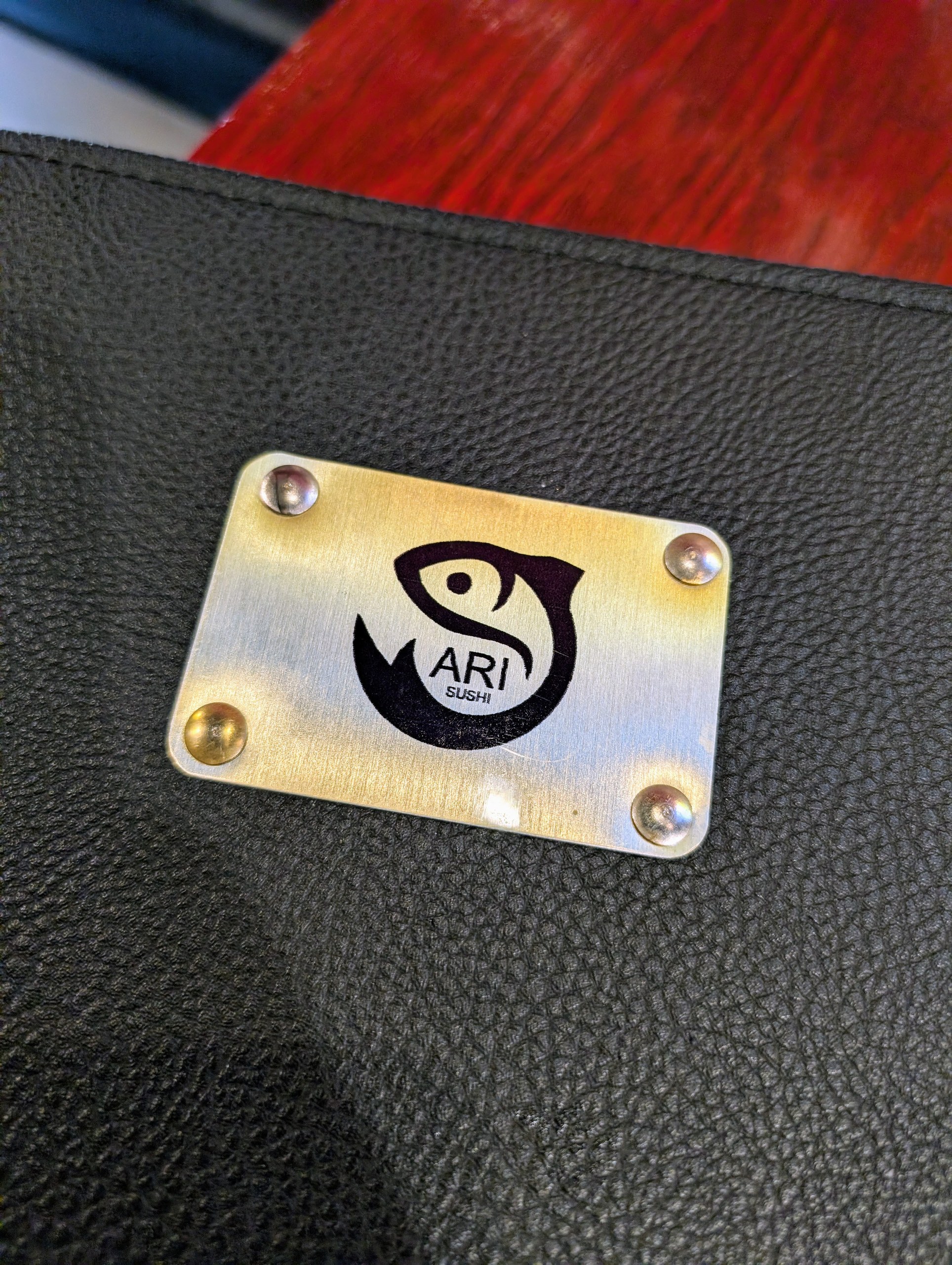 Brushed metal Ari Sushi logo plate inset into a black leather menu cover