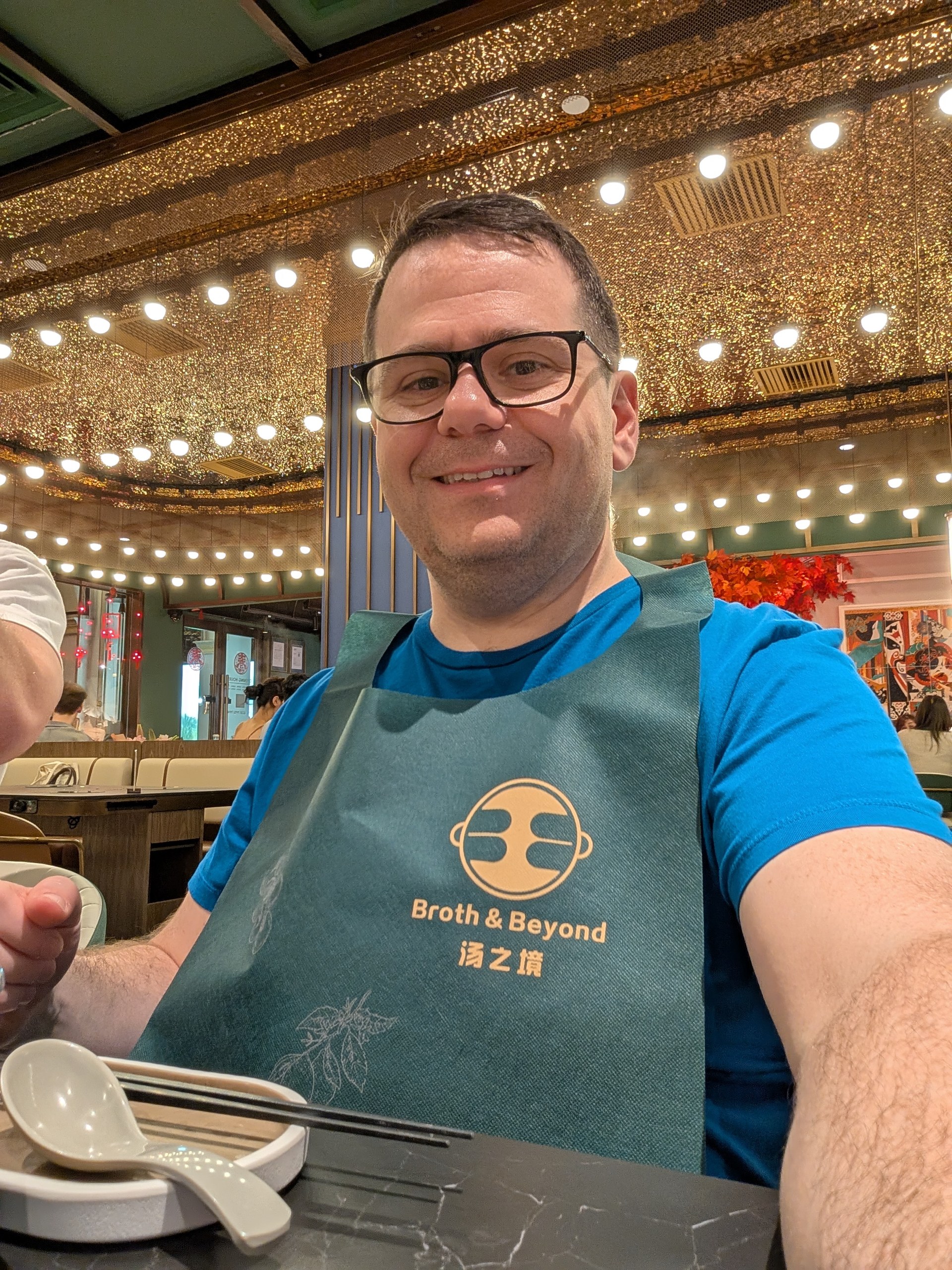 Alex at Broth and Beyond CHIJMES wearing the branded teal apron, chandelier-style ceiling behind him