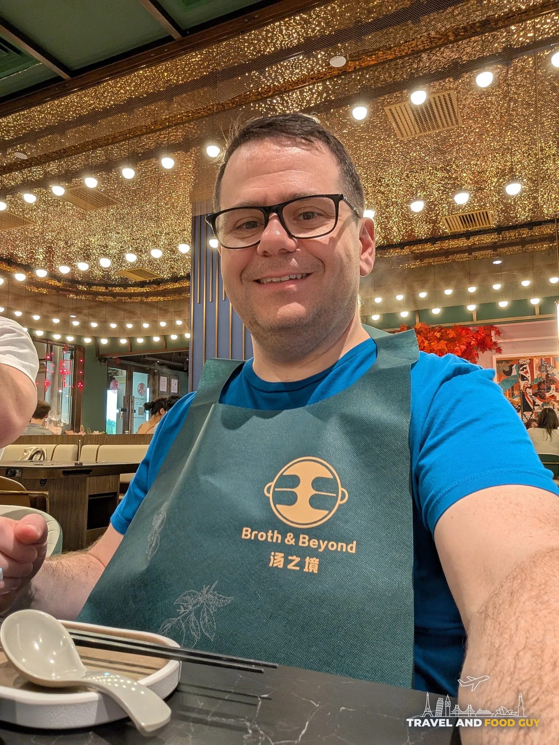 Alex at Broth and Beyond CHIJMES wearing the branded teal apron, chandelier-style ceiling behind him