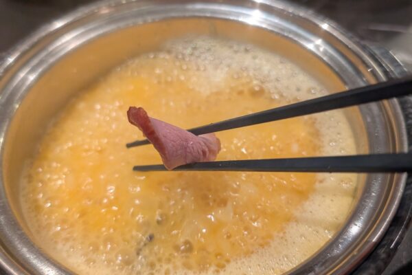 Chopsticks holding a slice of smoked meat over the bubbling Signature Golden Chicken Fungus broth at Broth and Beyond