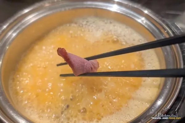 Chopsticks holding a slice of smoked meat over the bubbling Signature Golden Chicken Fungus broth at Broth and Beyond