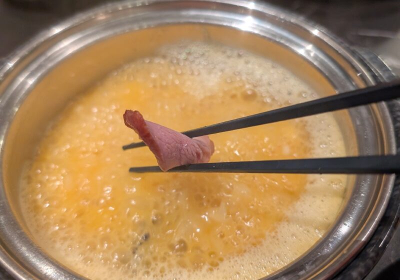 Chopsticks holding a slice of smoked meat over the bubbling Signature Golden Chicken Fungus broth at Broth and Beyond