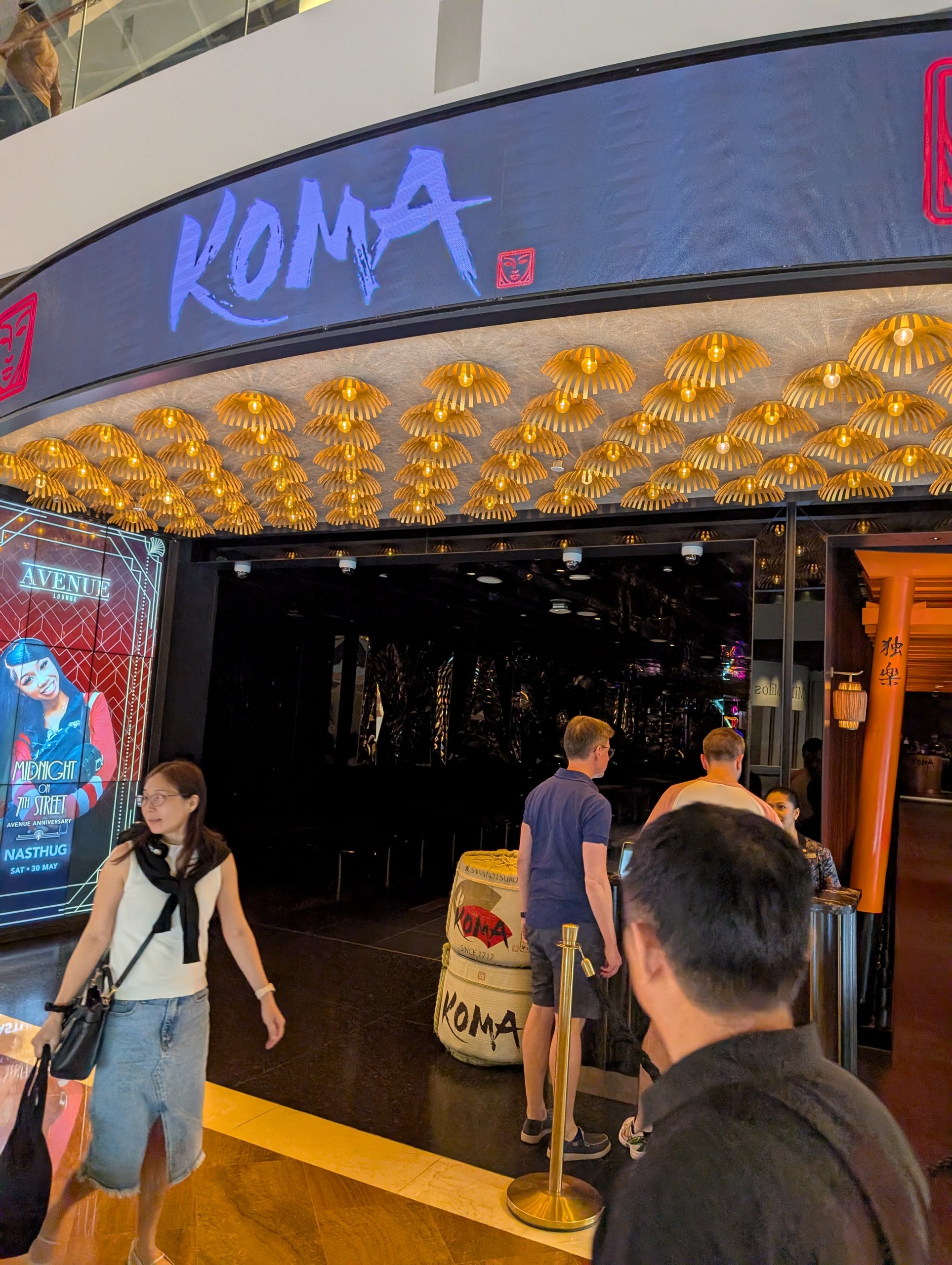 The entrance to KOMA Singapore at The Shoppes at Marina Bay Sands, with a curved canopy of gold paper parasols above a barrel marked KOMA.