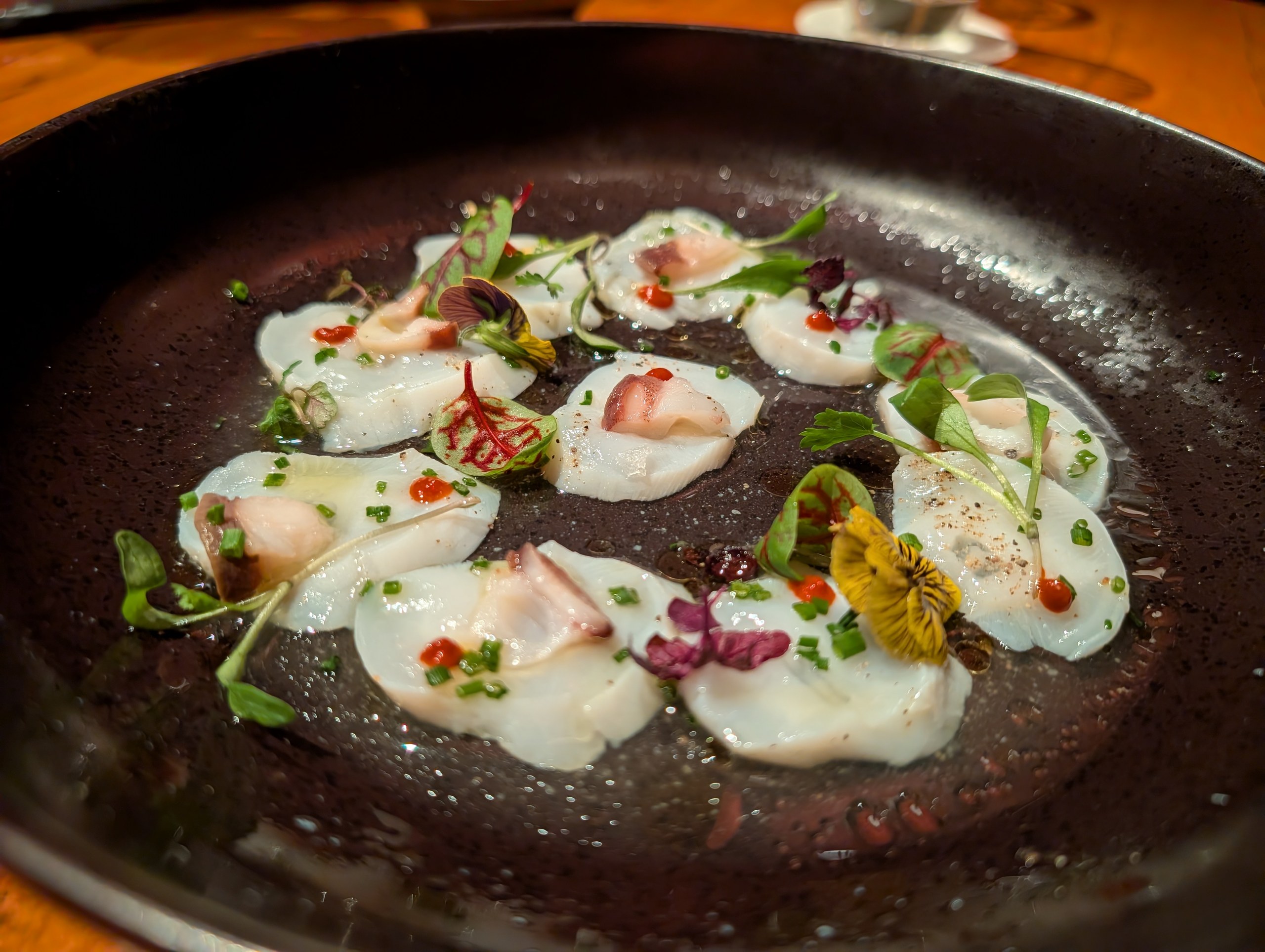 Hokkaido scallop sashimi at KOMA Singapore, paper thin slices in a dark pan with scallion oil and mustard seeds.