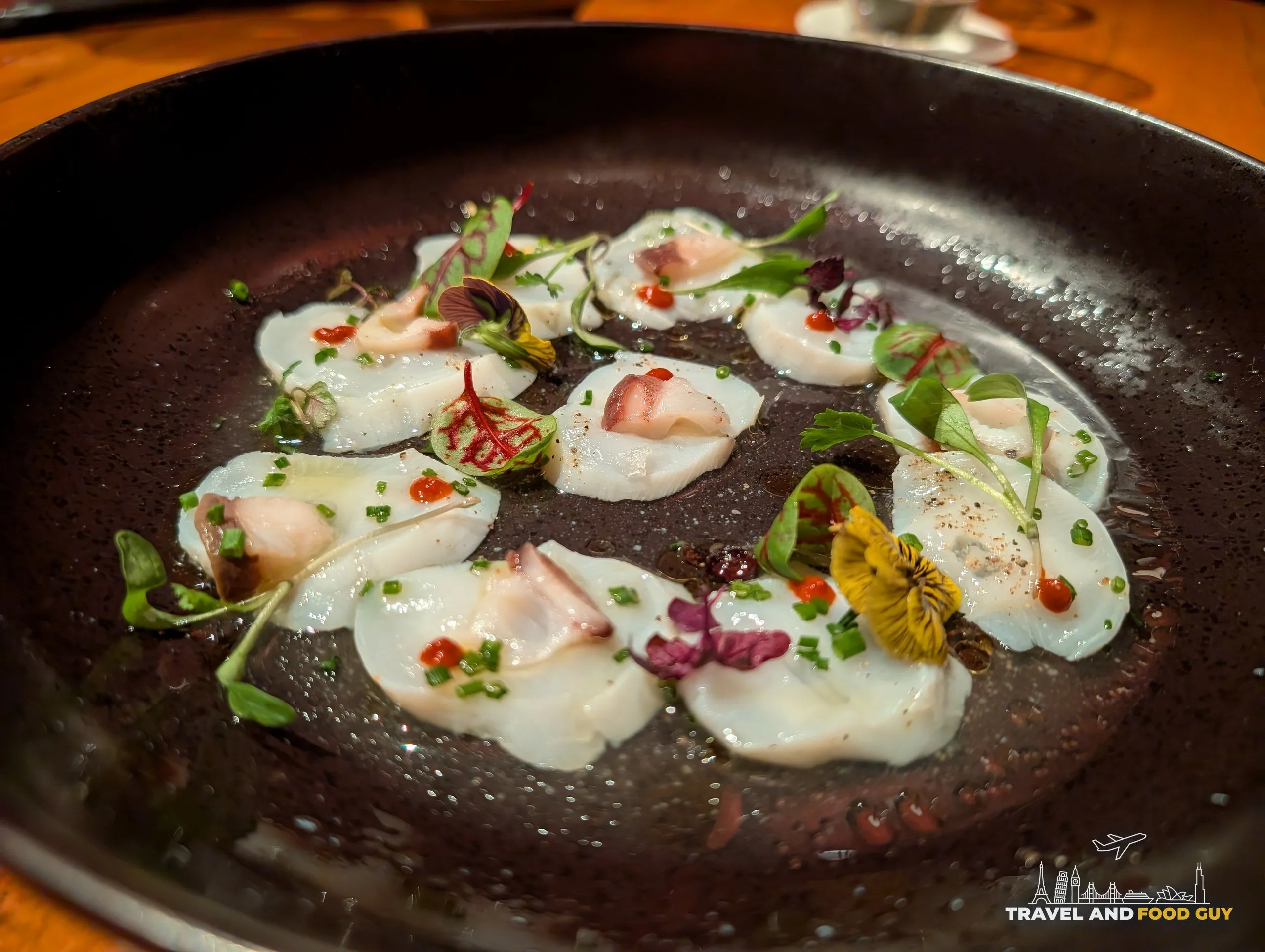 Hokkaido scallop sashimi at KOMA Singapore, paper thin slices in a dark pan with scallion oil and mustard seeds.
