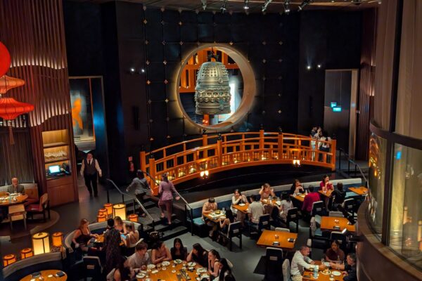 The KOMA Singapore dining room from the upper mezzanine, showing the bronze bell, the wooden bridge, and the paper parasol ceiling.