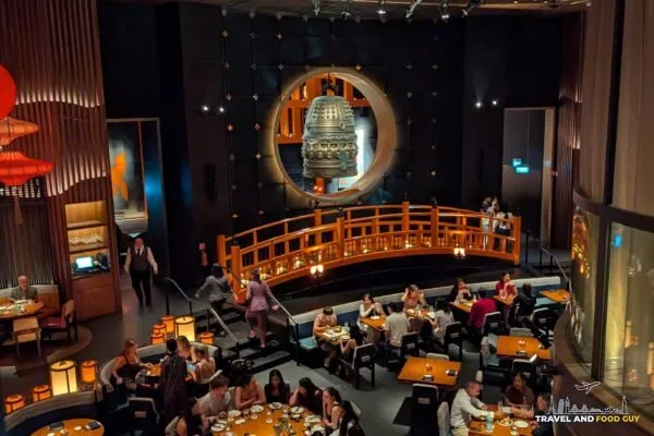 The KOMA Singapore dining room from the upper mezzanine, showing the bronze bell, the wooden bridge, and the paper parasol ceiling.