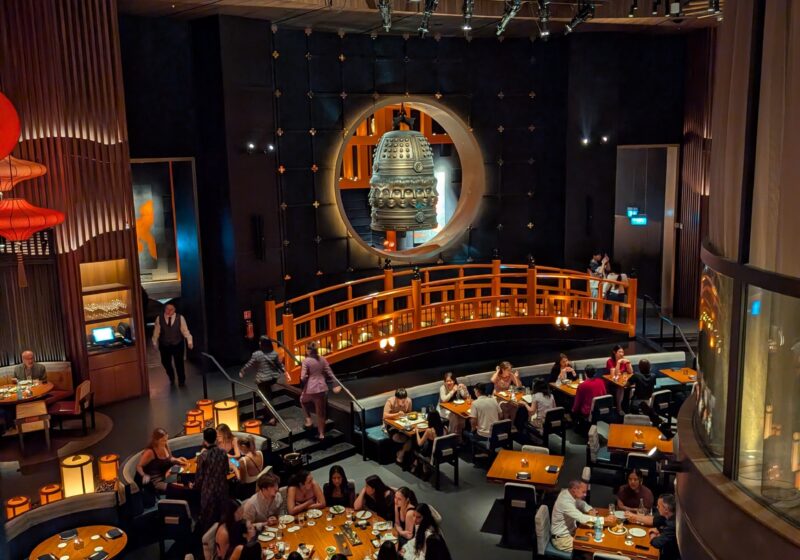 The KOMA Singapore dining room from the upper mezzanine, showing the bronze bell, the wooden bridge, and the paper parasol ceiling.
