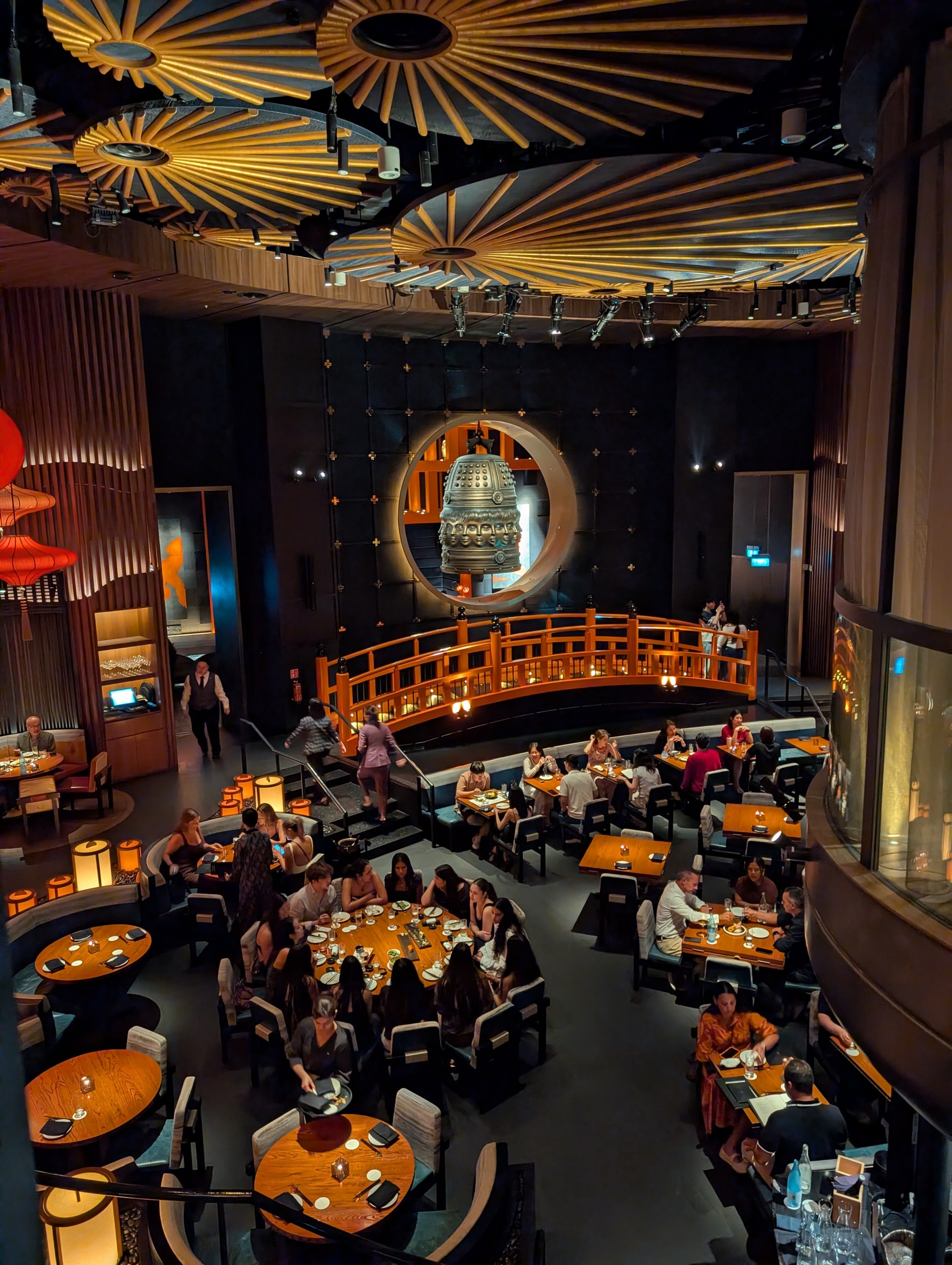 The upper mezzanine view of the KOMA Singapore dining room, showing the bronze bell, the wooden bridge, and the ceiling of paper parasols.